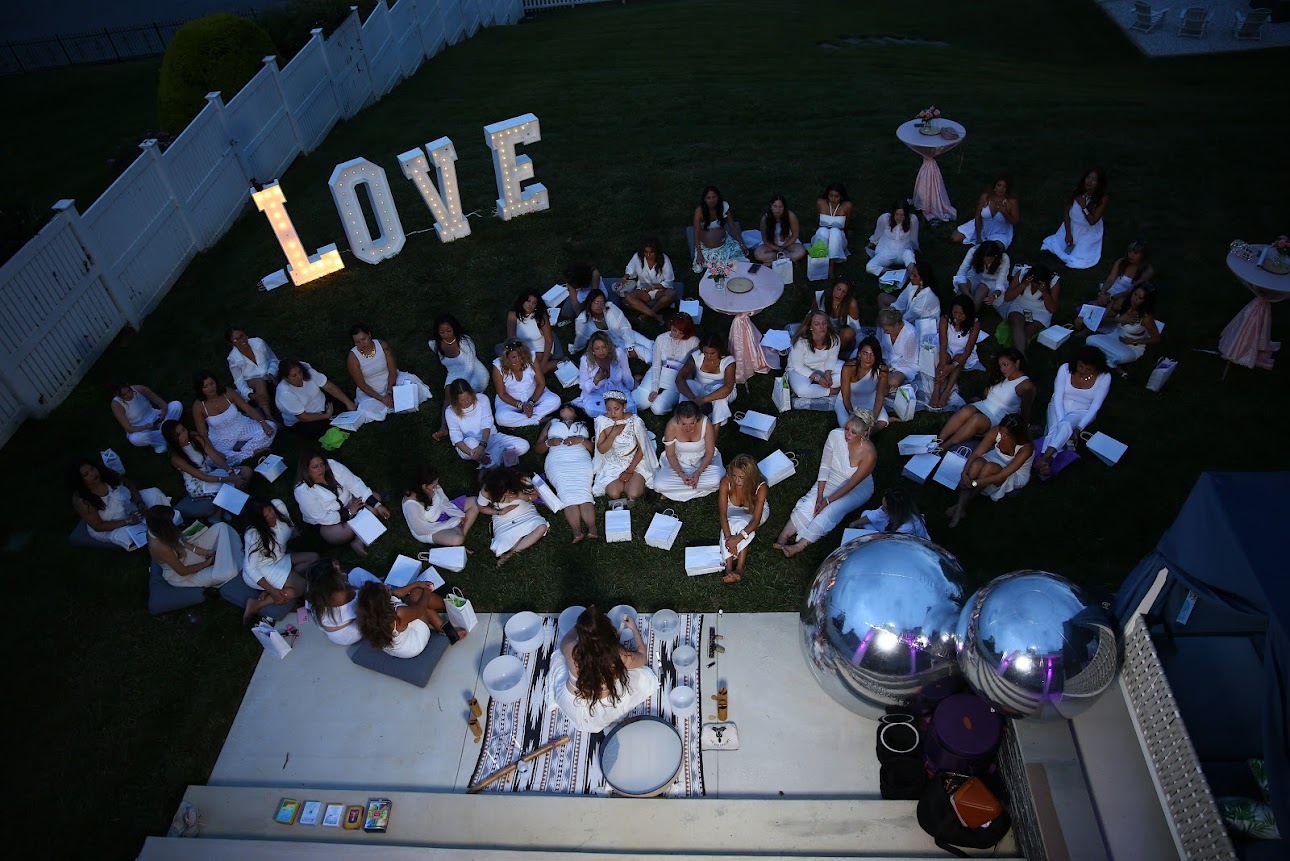 Women in white sitting on the grass beside a glowing LOVE sign, seen from above
